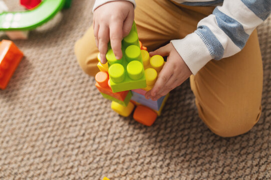 A 5-6-year-old boy plays with a colorful plastic construction set and blocks, building houses and cars on the carpeted floor at home alone. close-up of a hand, no face, unrecognizable
