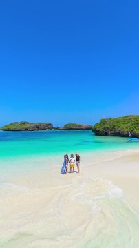 Aerial view of two young women and a young man standing on white sand near turquoise water and rocky islands at Watamu Beach, Kenya.