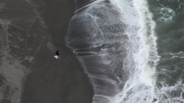 Aerial view of the black sands of Pandansari Beach meet the foamy, turquoise waves in a striking contrast of textures and tones with people playing alone, Bantul Regency, Indonesia.