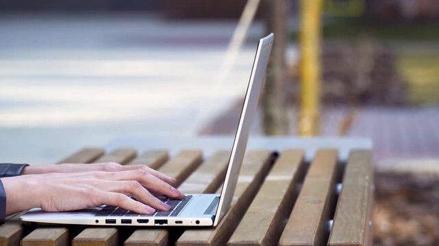 Female hands typing on laptop keyboard at wooden bench outdoors, copy space
