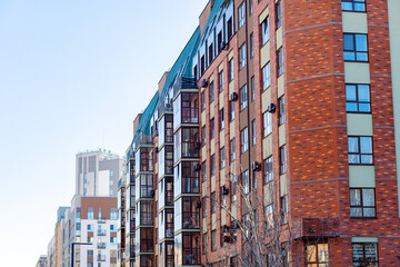 Fototapeta premium Bright red brick building rises beside blue sky. Windows line the facade, some with balconies. Air conditioners hang near every window. Street view shows urban architecture with clean lines
