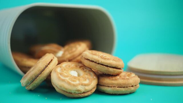 Biscuits with cream filling spilling out of fallen container over bright turquoise surface.