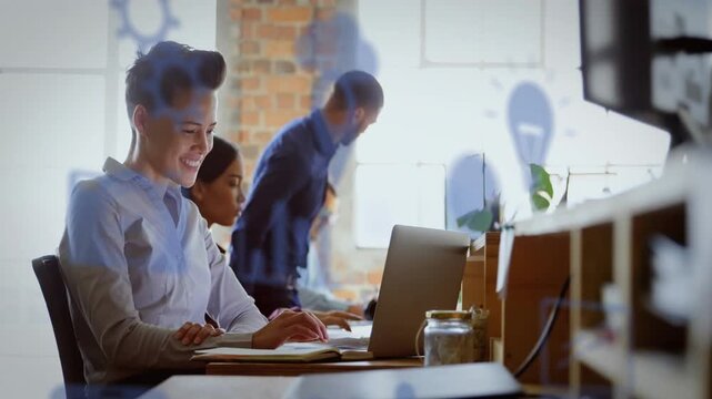 Typing short hair woman in light shirt referencing notebook at bench with laptops, blue icons