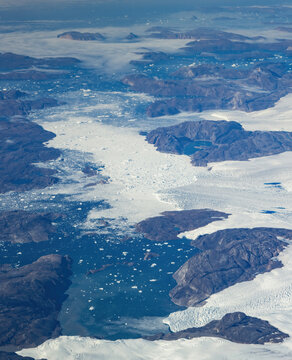 Aerial view of a glacial river dotted with icebergs flows through rugged, rocky terrain, contrasting with the vast, white expanse of ice, Narsarsuaq, Greenland.
