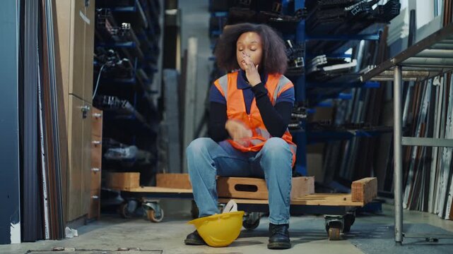 Fatigued African American female warehouse worker taking off a yellow hard hat and sitting down to rest after a hard workday.