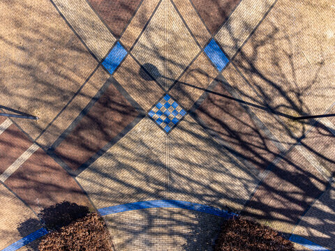 Aerial view of patterned pavement with diamond and square shapes in brown and blue hues, overlaid with the stark shadows of bare trees, Maastrichtkwartier, Flevoland, Netherlands.