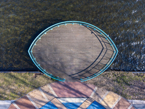 Aerial view of a curved wooden platform with a blue railing, overlooking the shimmering water and abstract patterns below, Maastrichtkwartier, Flevoland, Netherlands.