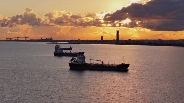 Aerial drone view of multiple oil tankers anchored near an industrial port at sunset with cranes and harbor infrastructure visible on the horizon, Limassol, Cyprus