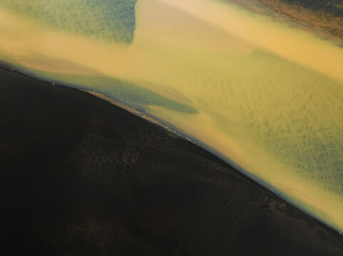 Aerial view of the glacial river's silty, ochre waters meeting the stark black sands, creating a mesmerizing contrast of color and texture, Iceland, Iceland.