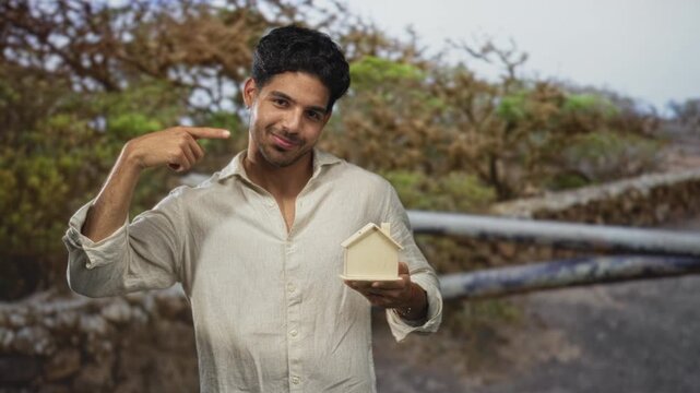Young man holds miniature wooden house and points finger to chest on a street; homeownership pride aspiration.