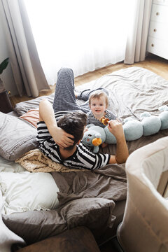 Happy baby and father playing on bed with soft and wooden toys in rustic bedroom. Family domestic life. Stylish cute dad and son having fun together, Happy Father's day