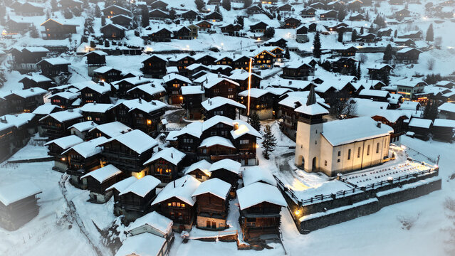 Aerial view of snow-dusted chalets nestled around a brightly lit church, creating a warm glow against the cool, crisp winter landscape, Grimentz, Switzerland.