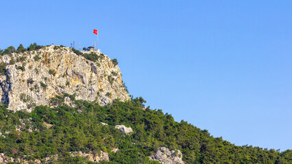 Naklejka premium Wide landscape view of Mount Calis (Idyros) in Kemer, Turkey with the Turkish flag on the summit and large blue sky copy space on a sunny day