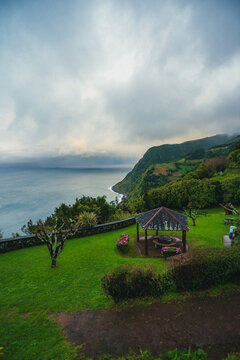 Aerial view of a rustic gazebo nestled on vibrant green grass overlooking the rugged coastline meeting the vast ocean under a cloudy sky, Nordeste, Azores, Portugal.
