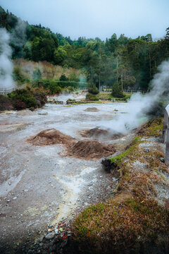 View of geothermal landscape with steaming vents and bubbling mud, a mix of earthy browns and vibrant greens under a hazy sky, Furnas, Azores, Portugal.