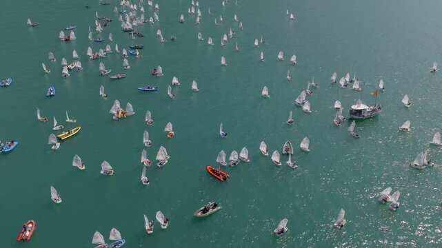 Cinematic orbit drone shot capturing a dynamic sailing regatta on Lake Garda, Italy. Multiple sailboats race across clear blue water as the drone smoothly circles the fleet, creating a dramatic scene