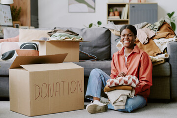 Portrait of young Black woman sorting folded clothes into donation boxes at home, supporting decluttering and charitable giving. Ideal for nonprofit campaigns, resale apps, sustainability marketing