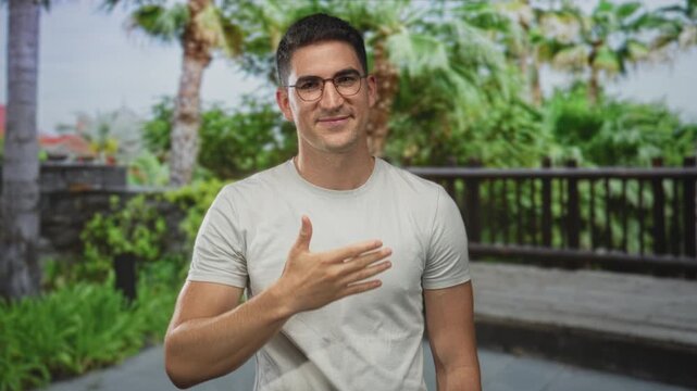 Man, young hispanic, wearing glasses and a white t shirt, placing hand on chest on a street outdoors near wooden railing; sincerity trust.