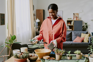 Young Black woman repotting houseplants at home, holding soil and planter while gardening. Ideal for home lifestyle, self care, indoor hobby, sustainability marketing