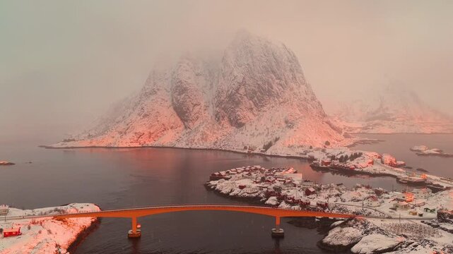 Cinematic aerial shot of the snow covered Lofoten archipelago in Norway during a misty pink sunset, featuring a bridge and traditional coastal village.