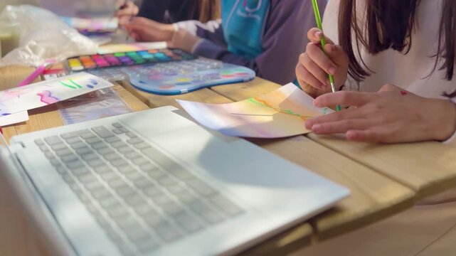 Children painting with watercolor at outdoor table, using laptop for inspiration. Creative learning, art activity, teamwork, education, colorful scene.
