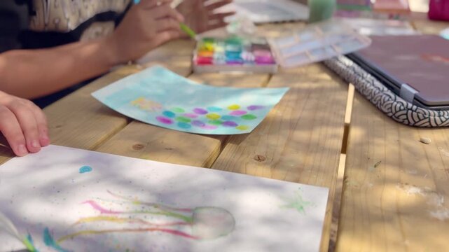 Children painting with watercolor at outdoor table, using laptop for inspiration. Creative learning, art activity, teamwork, education, colorful scene.