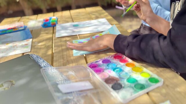Children painting with watercolor at outdoor table, using laptop for inspiration. Creative learning, art activity, teamwork, education, colorful scene.