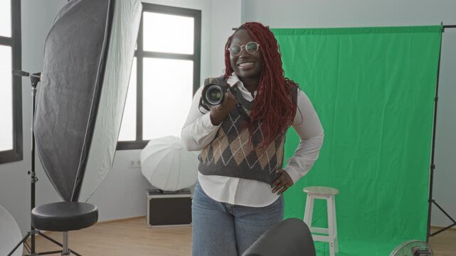 Woman holding camera and smiling, hand on hip, wearing glasses and argyle vest, standing by green screen and softbox in studio; creative confidence.