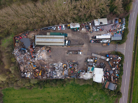 Aerial view of a recycling center where piles of scrap metal and crushed vehicles contrast starkly with the surrounding green fields and roadside, Stafford, England, United Kingdom.