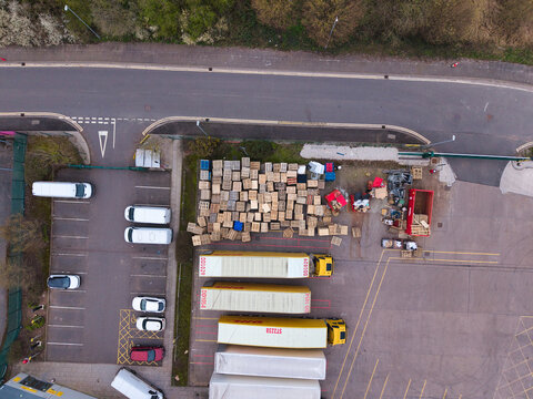 Aerial view of trucks and stacked boxes create a geometric pattern of yellow, brown, and grey, contrasting with the surrounding tarmac, Northampton, United Kingdom.