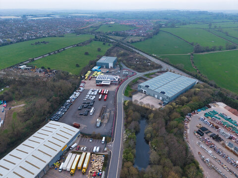 Aerial view of industrial buildings and vehicles nestled amongst lush green fields and tree lines, with a small pond reflecting the overcast sky, Redditch, United Kingdom.
