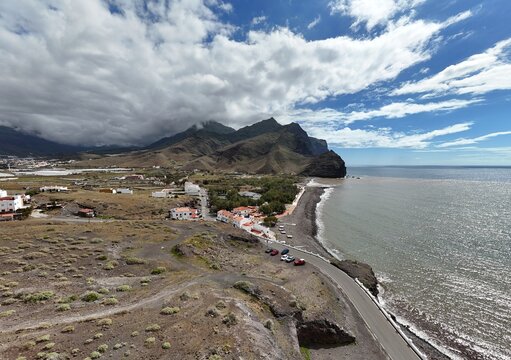 Aerial view of dark volcanic sands meeting the turquoise ocean, a rugged coastline contrasting with the distant mountains, Puerto de la Aldea, Canarias, Spain.