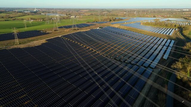 Aerial view of the expansive Tye Lane solar farm displaying dark panels contrasted against green fields and transmission towers, Ipswich, United Kingdom.