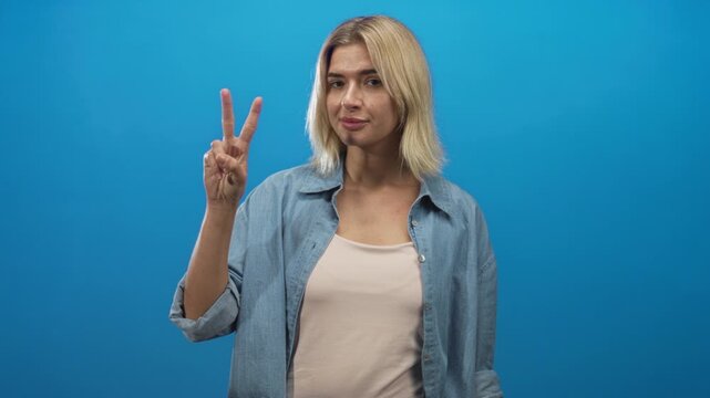 Woman holds up fingers counting, hand raised in studio with blue backdrop, denim shirt and beige top; gesture playful communication.