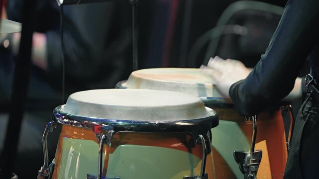 Close-up of a percussionist's hands in motion, playing conga drums on stage. The dynamic shot captures the rhythm and energy of a live jazz concert with focused lighting and a dark background
