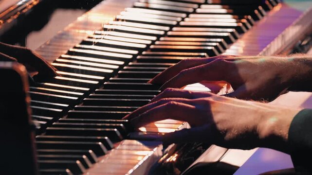 Close up a pianist's hands in motion on a grand piano keyboard. Moody purple and orange lighting highlights the musician during a live jazz concert performance