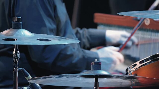 Close-up of drum cymbals with a blurred musician playing in the background. Cool, atmospheric lighting highlights the metallic texture, capturing the energy of a live jazz concert or music festival