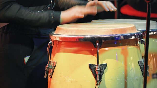 Close up of a percussionist's hands playing conga drums. Vibrant stage lighting creates a warm, energetic mood at a live music concert or festival. Focus on the traditional instrument
