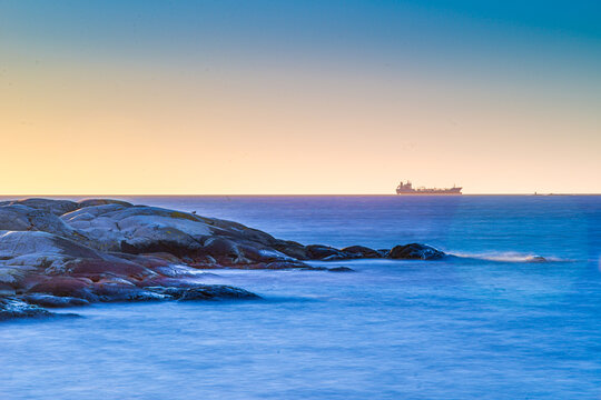 Distant cargo ship on calm ocean horizon with rocky coastal foreground.