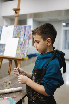Young boy paints with focus in art studio wearing smock surrounded by colorful canvases and art supplies