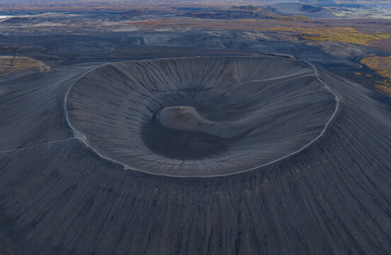 Aerial view of the stark, grey-black volcanic crater, its textured slopes contrasting with the distant autumn-tinged vegetation, shapes a surreal landscape, Myvatn, Thingeyjarsveit, Iceland.