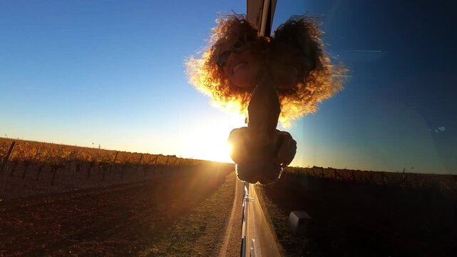 Woman enjoying travel experience from camper window at sunset, feeling wind and motion on open road. Passenger side view with moving landscape, countryside and summer evening atmosphere