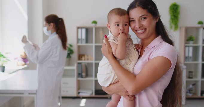 Smiling mother holds baby at medical clinic as a pediatrician and doctor get ready for immunization holds syringe for injection. Mother holds child before vaccination. Healthcare and medicine