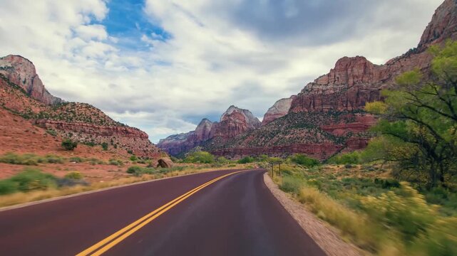 wide-angle view from a moving vehicle captures the majestic landscape of Zion National Park in Utah. The winding road leads into a canyon surrounded by towering, reddish-orange sandstone cliffs and ro