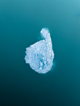 Aerial view of a solitary, sculpted iceberg adrift in the glacial lagoon, its icy blue form contrasting with the dark, placid waters, Jokulsarlon, Iceland.