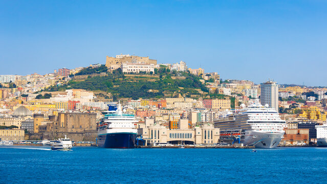 Panoramic view of Naples Italy coastline with harbor, cruise ships and historic buildings. Naples features Castel Sant Elmo on hill above urban architecture. Clear weather, calm sea, bright daylight 