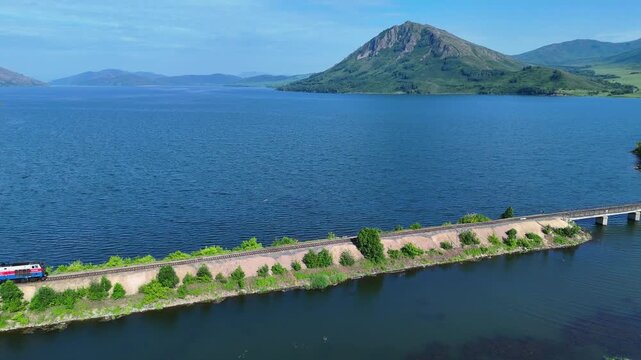 Cinematic drone footage of a train traveling along a narrow strip of land surrounded by the waters of the Bukhtarma Reservoir. On a clear sunny day, the shot features the calm water surface