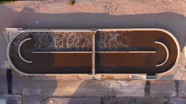 Top-down view of an oval aeration basin at a wastewater treatment plant with active surface agitation and brown activated sludge