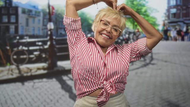 Senior hispanic woman forms heart with hands above head, tied red striped shirt and slight bare midriff on cobblestone street; joyful travel.