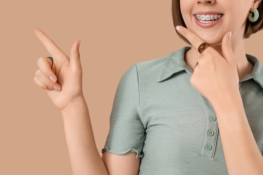 Young woman with dental braces pointing at something on beige background, closeup